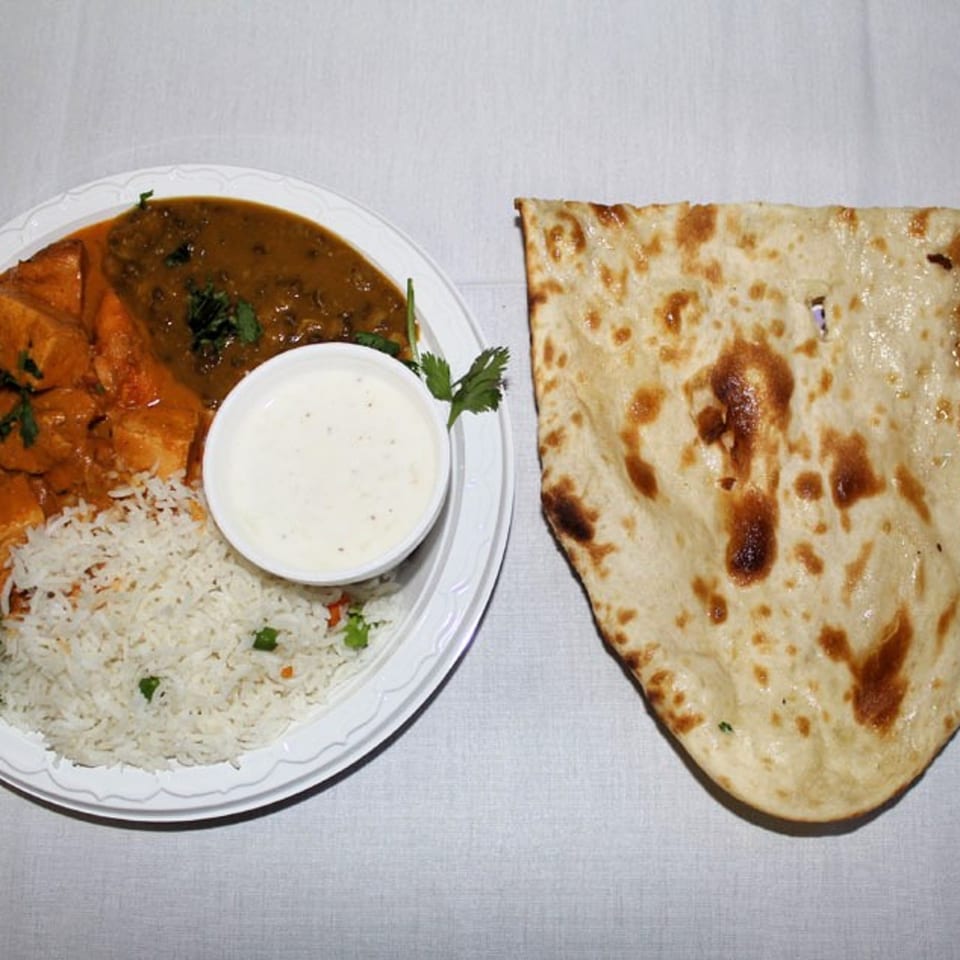 A plate of Indian food with rice, two curries, raita, and a side of naan bread.