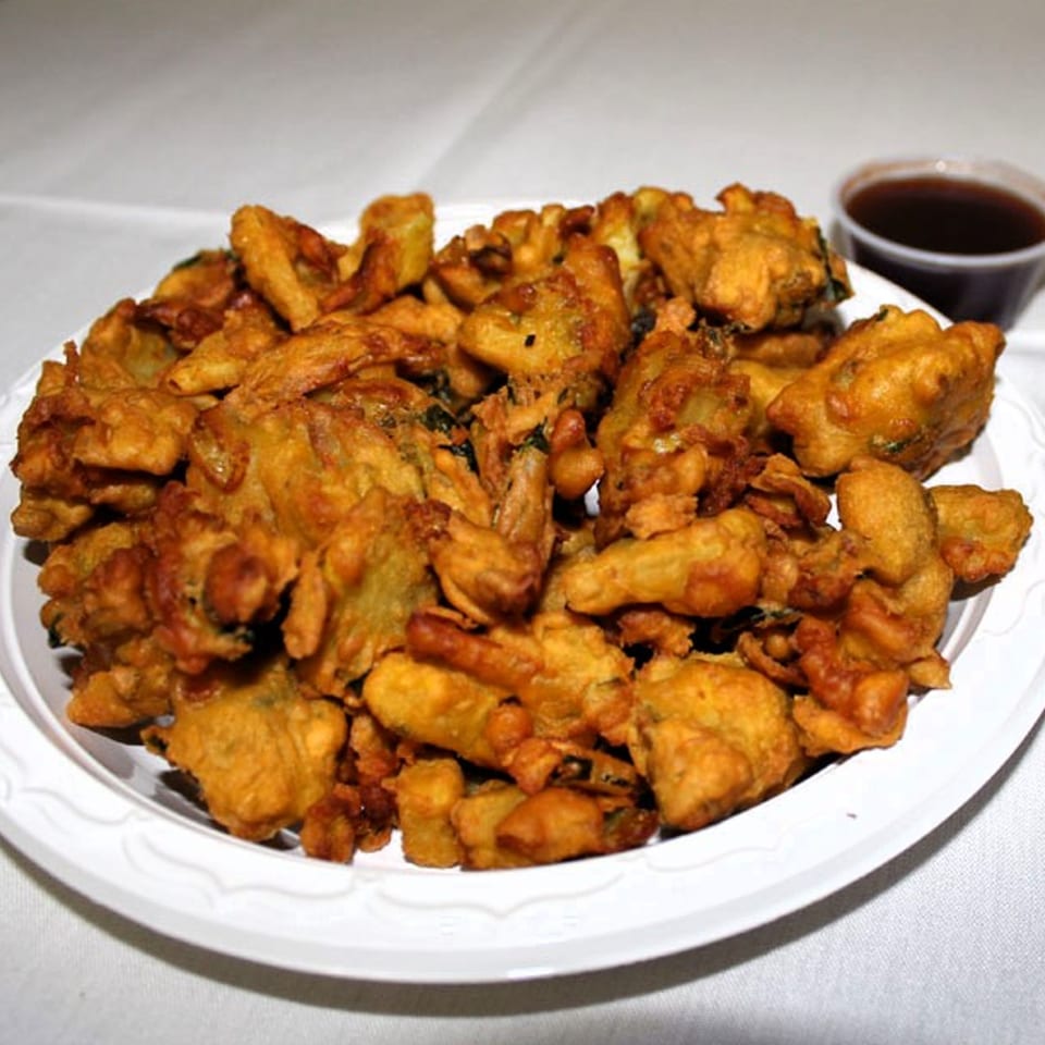 A white plate piled high with golden-brown, crispy Indian pakoras, with a small cup of dipping sauce.