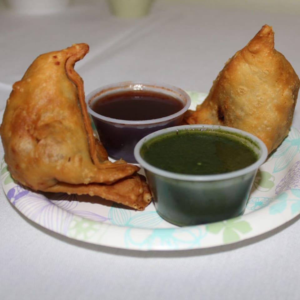 Two golden-brown Indian samosas with two small cups of dipping sauces on a paper plate.